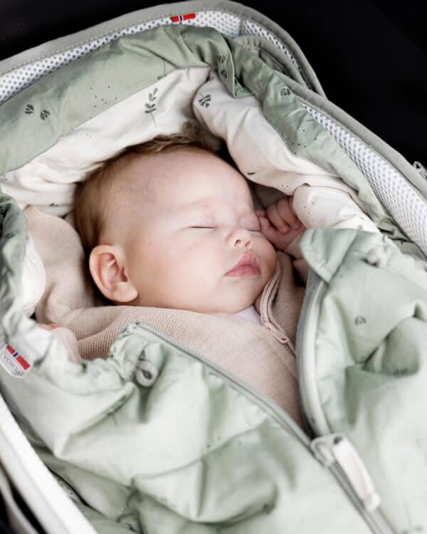 Gros plan d’un bébé endormi dans le Voksi Carry Me vert brume, intérieur crème.