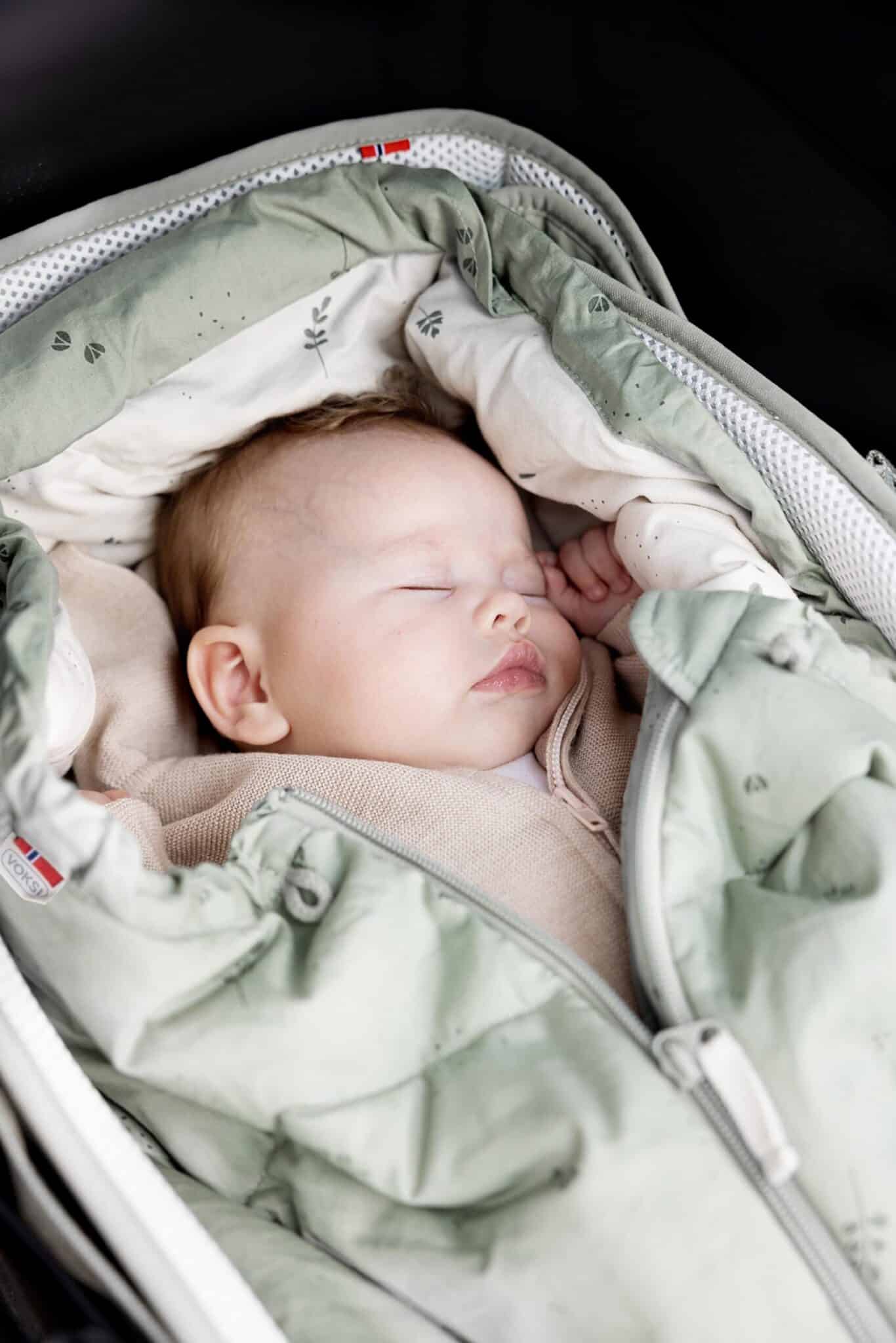 Gros plan d’un bébé endormi dans le Voksi Carry Me vert brume, intérieur crème.