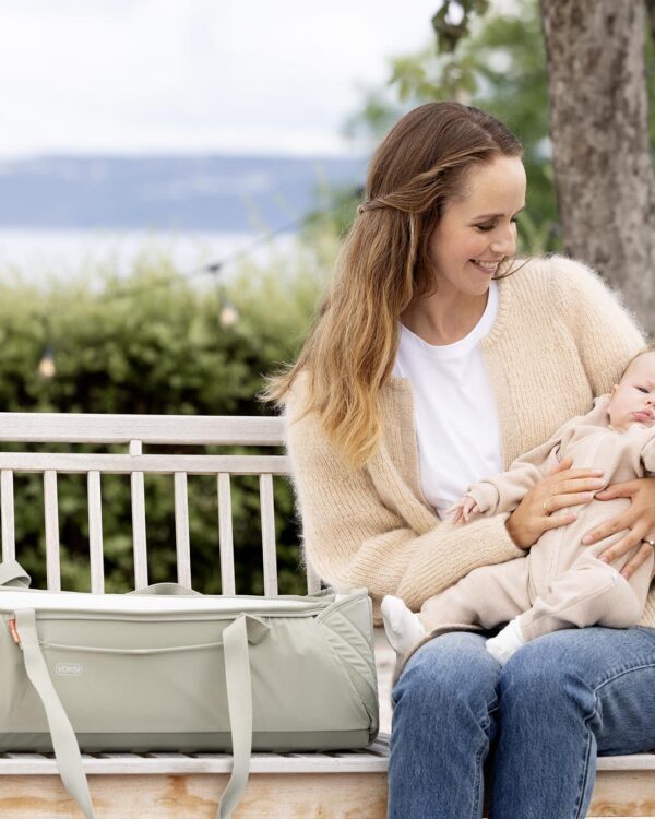 Maman avec bébé sur un banc, Voksi Carry Me Misty Green posée à côté, couffin nomade pour sorties.