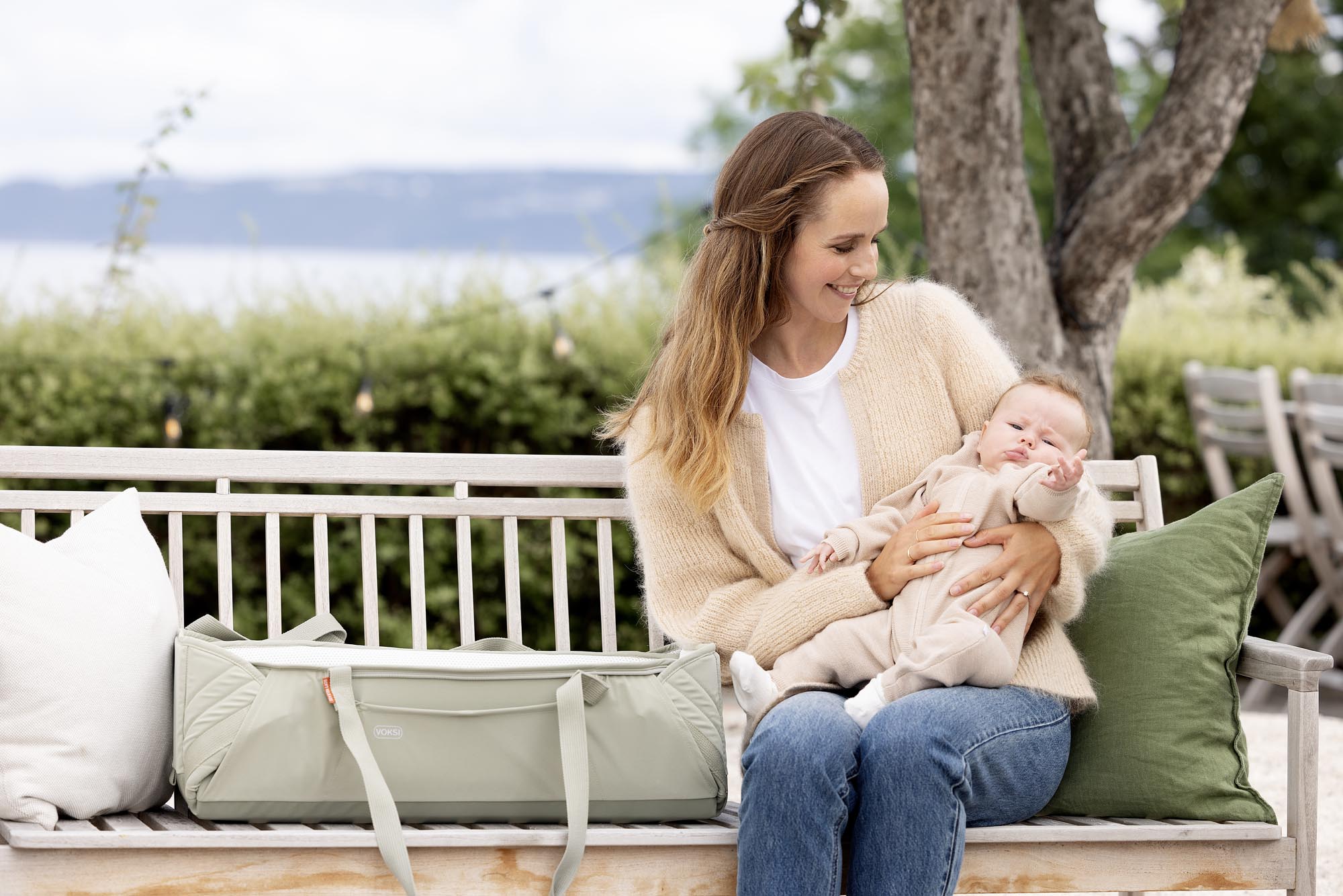 Maman avec bébé sur un banc, Voksi Carry Me Misty Green posée à côté, couffin nomade pour sorties.