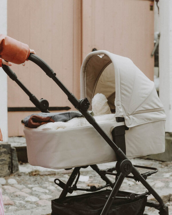 Maman poussant la nacelle Elodie MONDO coloris Moonshell sur châssis de poussette noire dans une rue pavée