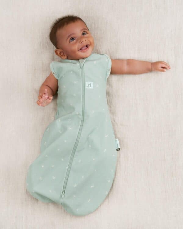 Bébé souriant portant le cocon d’emmaillotage ergoPouch Cocoon Sage vert d’eau, allongé sur fond beige