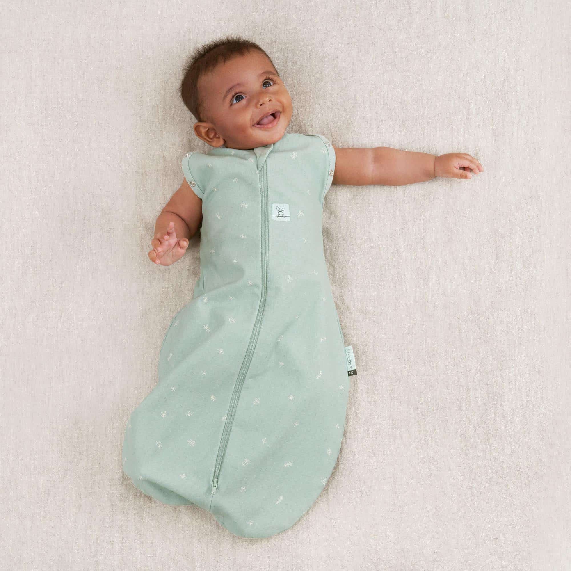 Bébé souriant portant le cocon d’emmaillotage ergoPouch Cocoon Sage vert d’eau, allongé sur fond beige