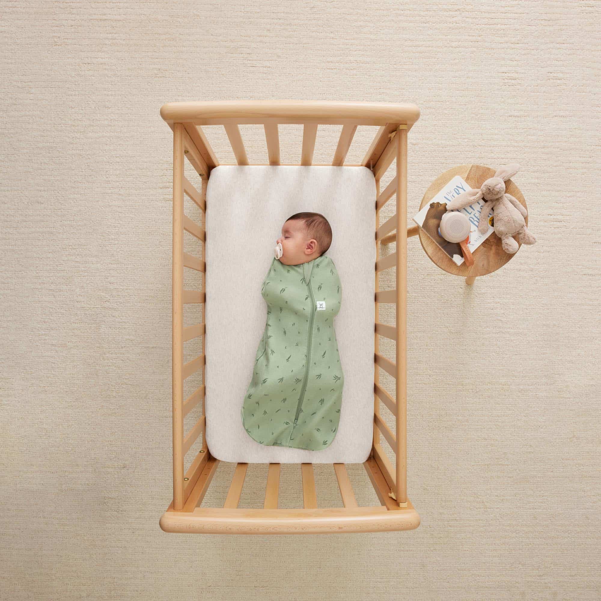 Bébé dans un lit en bois portant le cocon d’emmaillotage ergoPouch Cocoon Willow vert feuillage, vue du dessus